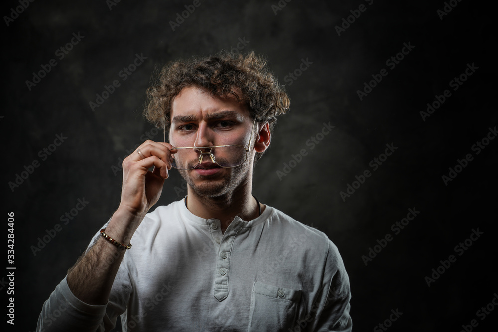 Grumpy looking young adult caucasian man posing in a dark studio on a grey background, wearing a white casual shirt and glasses Grumpy looking young adult caucasian man posing in a dark studio on a grey background, wearing a white casual shirt and glasses