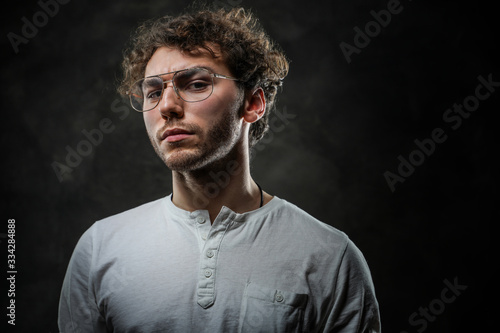 Thoughtful young student posing in a dark studio on a grey background, looking focused while wearing casual white shirt and glasses.