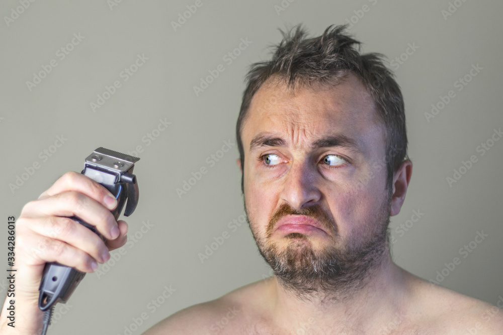 Portrait of a hairy man with unshaven face holding an electrical hair clipper and looks on it suspiciously. Portrait of a hairy man with unshaven face holding an electrical hair clipper and looks on it suspiciously.