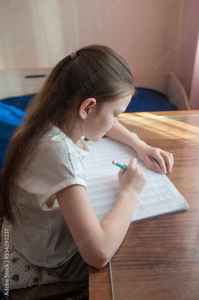 A school-age girl is studying at home at the table. Solves examples in a notebook. Stay at home. Coronovirus. A school-age girl is studying at home at the table. Solves examples in a notebook. Stay at home. Coronovirus.