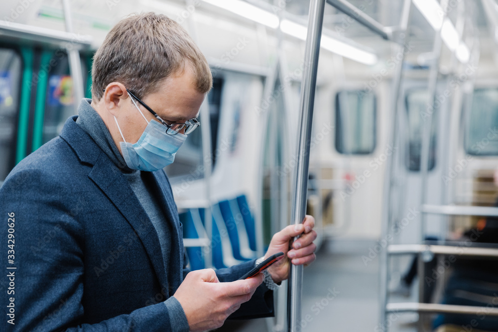 Fototapeta premium Serious man commutes to work in underground wears virus protective mask being always in touch with modern cellular poses in empty subway or metro. Distance concept. Public transport during coronavirus