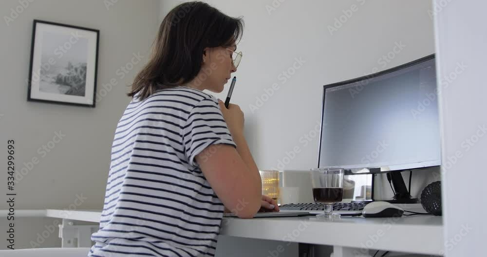Attractive young woman enjoying what she is doing as she works at computer in her beautiful home office. Female designer in eye glasses with reflection of screen lights reading news using PC computer Attractive young woman enjoying what she is doing as she works at computer in her beautiful home office. Female designer in eye glasses with reflection of screen lights reading news using PC computer