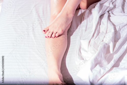 Bare feet of a girl with painted nails with red varnish on a white sheet in bed in bright sunshine