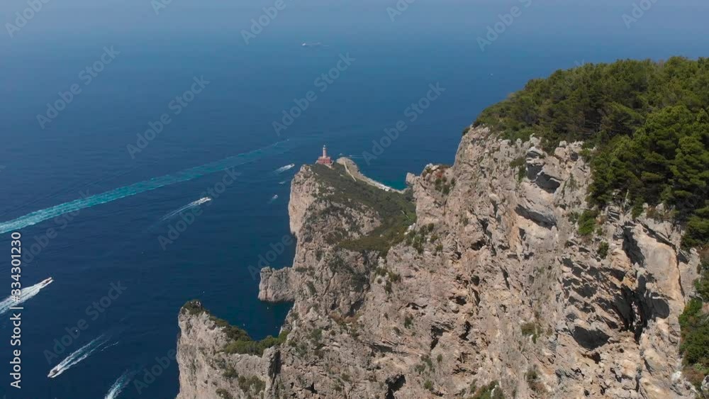 Aerial drone track forward top view of a stunning path hill with colorful trees toward a crowd of boats sailing in crystal clear seawater and Punta Carena lighthouse Capri