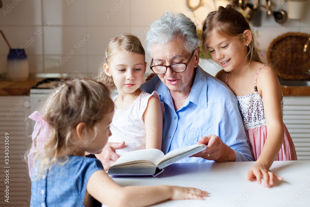 Reading book at home. Grandmother reads fairy tale for sisters. Kids listen to granny story in kitchen. Happy family leisure without gadgets. Children and senior woman togetherness. Lifestyle moment. Reading book at home. Grandmother reads fairy tale for sisters. Kids listen to granny story in kitchen. Happy family leisure without gadgets. Children and senior woman togetherness. Lifestyle moment.