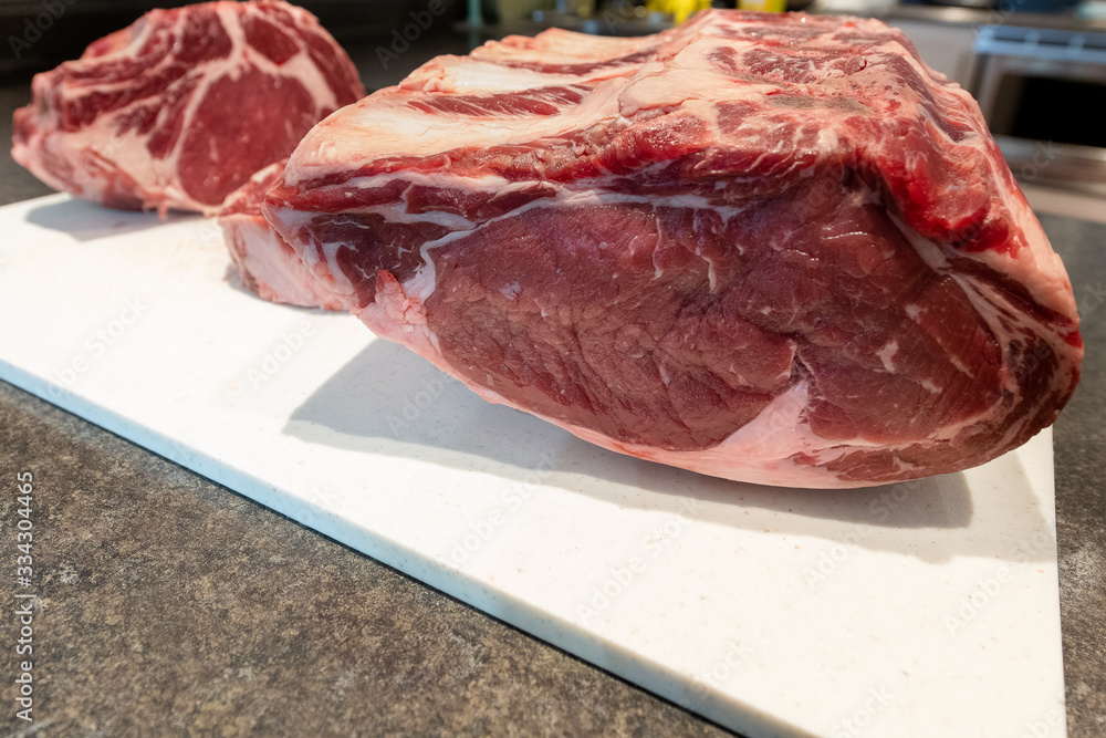Two large raw cuts of roast beef. The prime beef roasts have bones or ribs along the edge, fat for marbling and red juicy meat. The roasts are laying on a white plastic cutting board in a restaurant. 