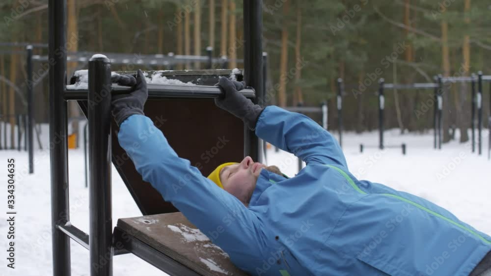 Medium shot of handsome man in blue windbreaker and yellow hat lying down on outdoor gym bench and doing leg raises while training in winter Medium shot of handsome man in blue windbreaker and yellow hat lying down on outdoor gym bench and doing leg raises while training in winter