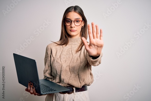 Young beautiful brunette woman working using laptop over isolated white background with open hand doing stop sign with serious and confident expression, defense gesture