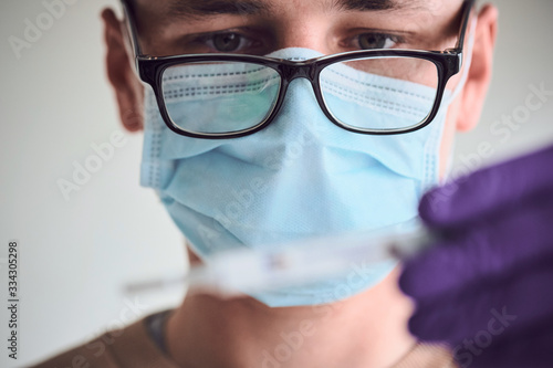 Young man wearing glasses, medical mask and disposable gloves looking at thermometer.