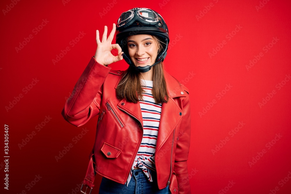 Young beautiful brunette motocyclist woman wearing motorcycle helmet and red jacket smiling positive doing ok sign with hand and fingers. Successful expression. Young beautiful brunette motocyclist woman wearing motorcycle helmet and red jacket smiling positive doing ok sign with hand and fingers. Successful expression.