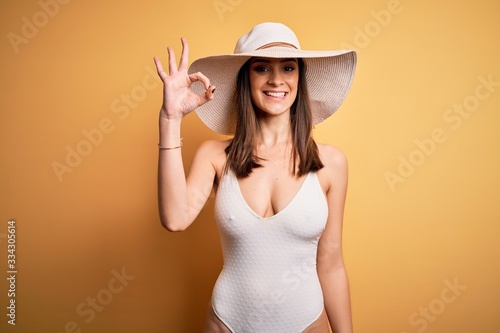 Young beautiful brunette woman on vacation wearing swimsuit and summer hat smiling positive doing ok sign with hand and fingers. Successful expression.