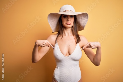 Young beautiful brunette woman on vacation wearing swimsuit and summer hat Pointing down looking sad and upset, indicating direction with fingers, unhappy and depressed.