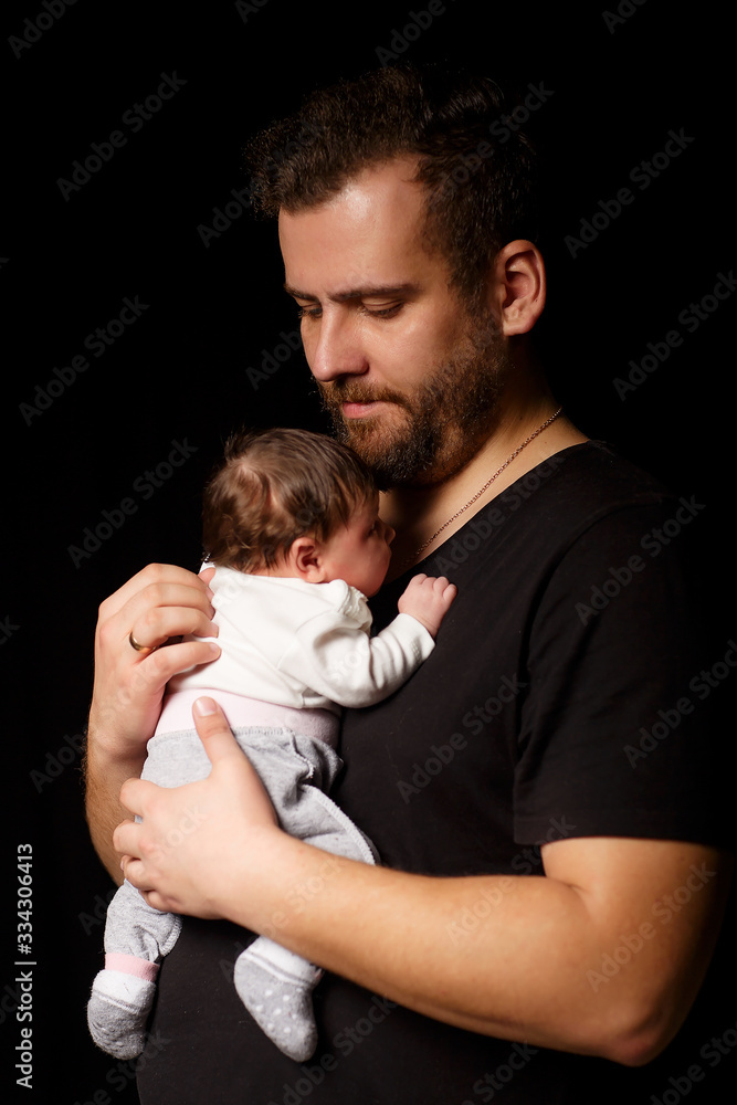 a bearded father in a black t-shirt holds a newborn baby. Dad kisses and hugs daughter or son on a black background a bearded father in a black t-shirt holds a newborn baby. Dad kisses and hugs daughter or son on a black background