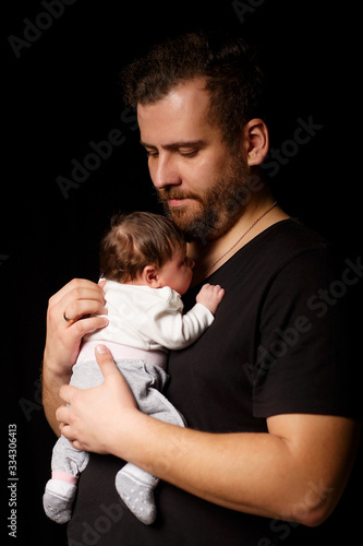 a bearded father in a black t-shirt holds a newborn baby. Dad kisses and hugs daughter or son on a black background