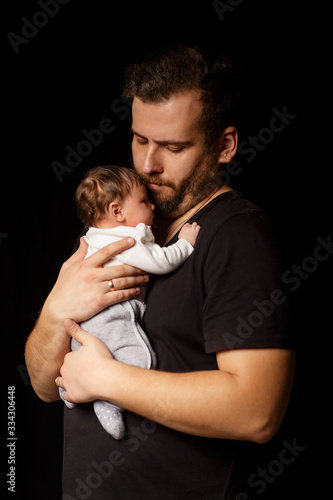 a bearded father in a black t-shirt holds a newborn baby. Dad kisses and hugs daughter or son on a black background