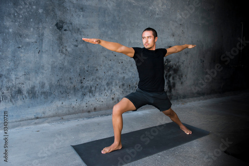 Man practicing yoga in front of a dark background. Pose is warrior II, Virabhadrasana. 