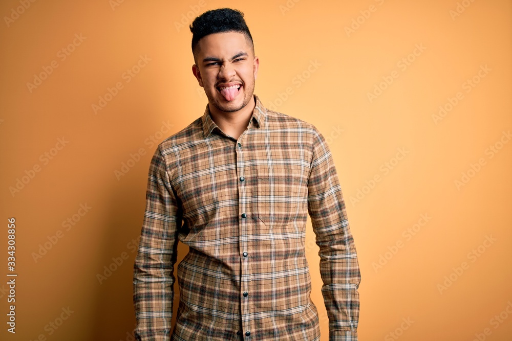 Young handsome man wearing casual shirt standing over isolated yellow background sticking tongue out happy with funny expression. Emotion concept. Young handsome man wearing casual shirt standing over isolated yellow background sticking tongue out happy with funny expression. Emotion concept.