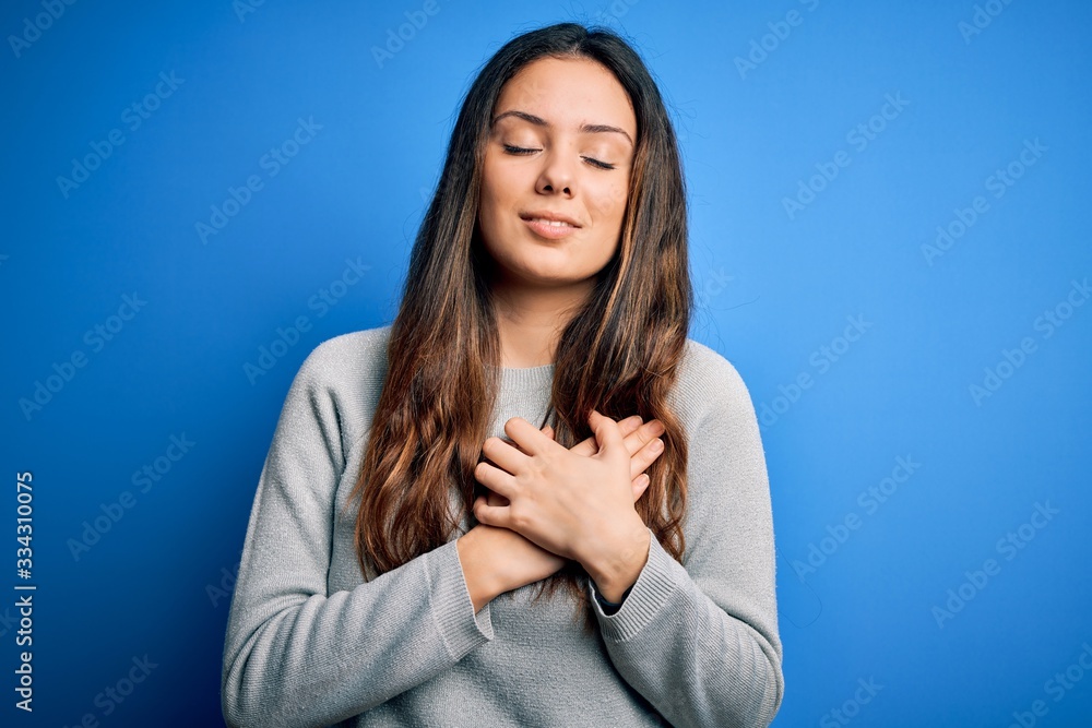 Young beautiful brunette woman wearing casual sweater standing over blue background smiling with hands on chest with closed eyes and grateful gesture on face. Health concept. Young beautiful brunette woman wearing casual sweater standing over blue background smiling with hands on chest with closed eyes and grateful gesture on face. Health concept.