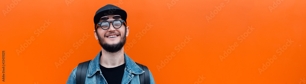 Panoramic banner, portrait of smiling millennial hipster guy wearing denim jacket, cap, backpack and glasses isolated on background of lush lava color with copy space. Panoramic banner, portrait of smiling millennial hipster guy wearing denim jacket, cap, backpack and glasses isolated on background of lush lava color with copy space.