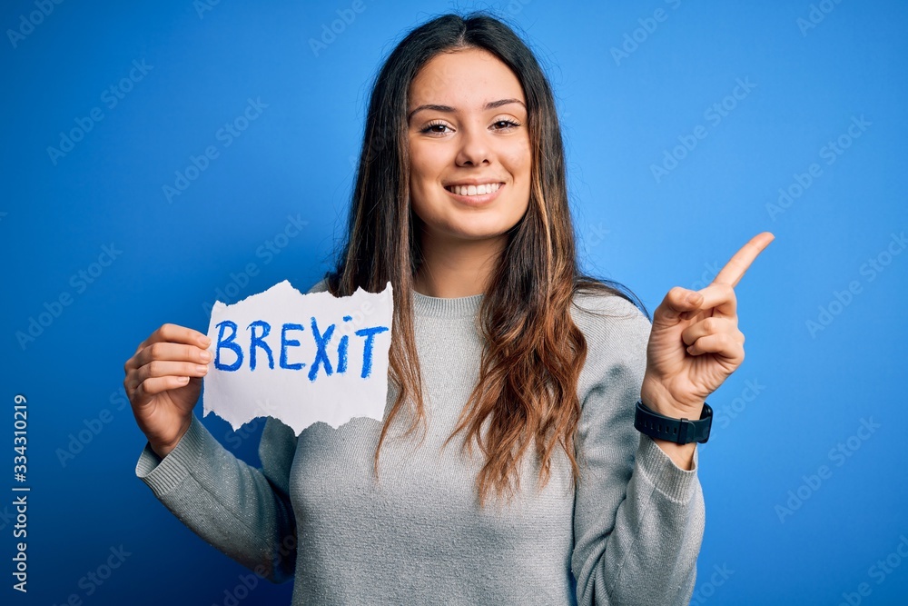 Young beautiful brunette woman holding paper with brexit message over blue background very happy pointing with hand and finger to the side Young beautiful brunette woman holding paper with brexit message over blue background very happy pointing with hand and finger to the side
