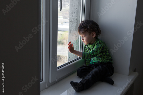 quarantine. a little boy sits in a window-sill and looks out the window bored. longing for fresh air and walks along the street. Forced home during quarantine due to the coronavirus pandemic