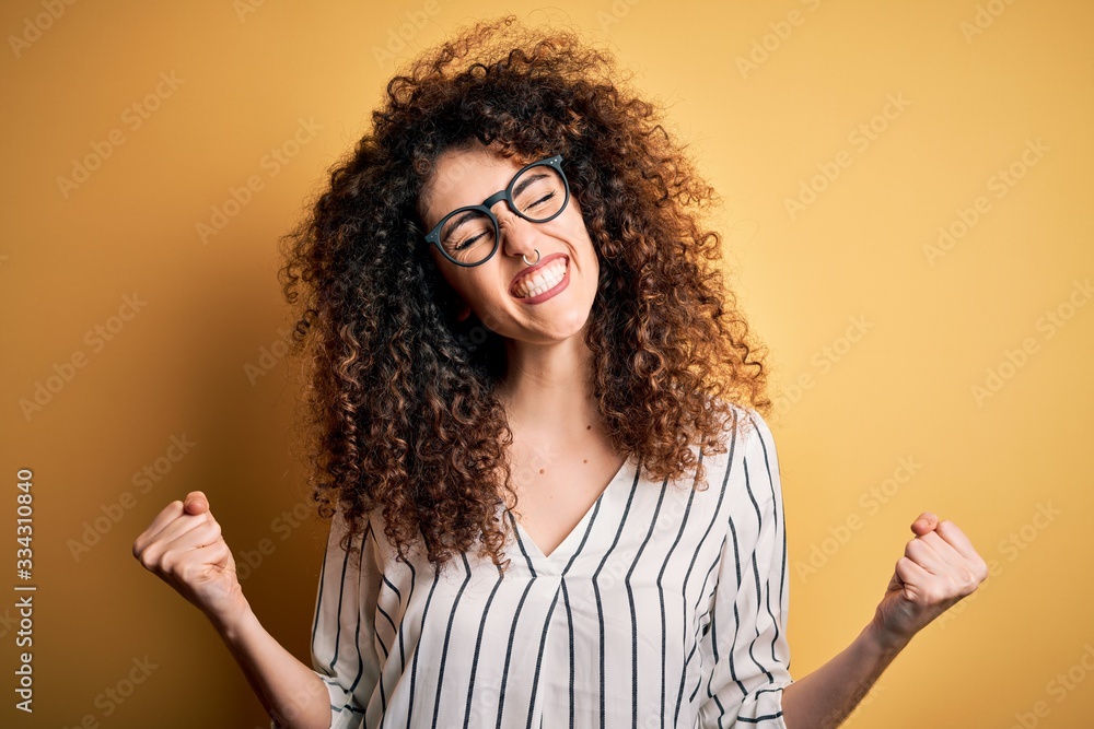 Young beautiful woman with curly hair and piercing wearing striped shirt and glasses very happy and excited doing winner gesture with arms raised, smiling and screaming for success. Celebration Young beautiful woman with curly hair and piercing wearing striped shirt and glasses very happy and excited doing winner gesture with arms raised, smiling and screaming for success. Celebration