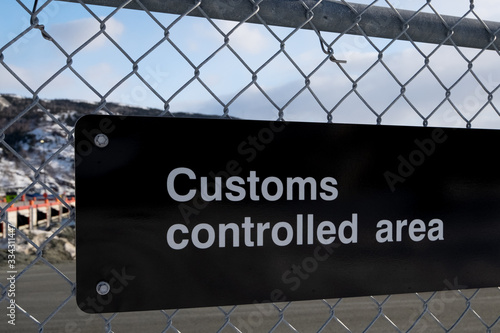 Customs controlled area warning sign, black and white in color. The sign is affixed to a metal chain link fence near a shipyard. The letters are white and the background of the sign is black. 