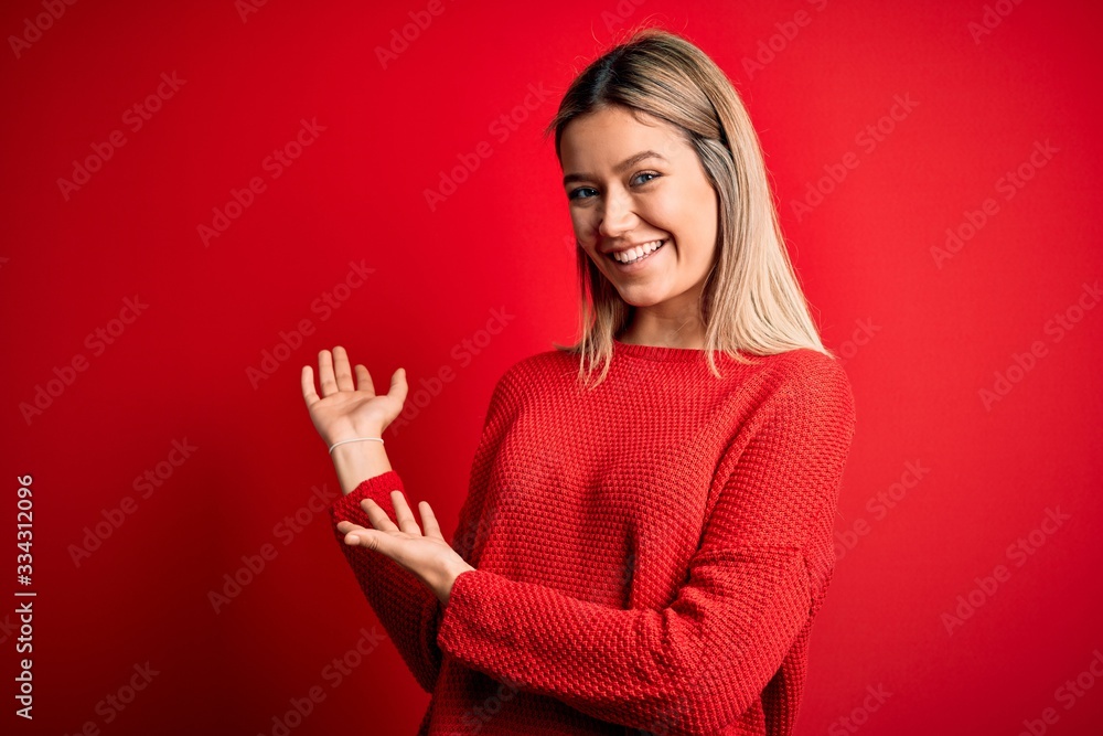 Young beautiful blonde woman wearing casual sweater over red isolated background Inviting to enter smiling natural with open hand Young beautiful blonde woman wearing casual sweater over red isolated background Inviting to enter smiling natural with open hand