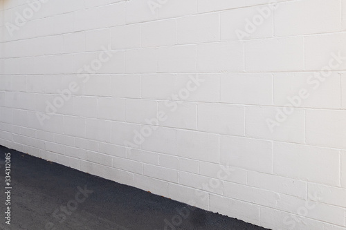 An exterior brick wall with a paved ground in an alley. The wall is composed of large concrete blocks.  The bricks have been painted a light grey color. The wall is on an angle to the ground.  