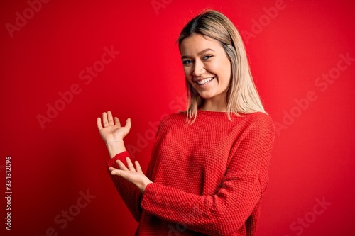 Young beautiful blonde woman wearing casual sweater over red isolated background Inviting to enter smiling natural with open hand