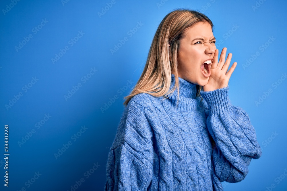 Young beautiful blonde woman wearing winter wool sweater over blue isolated background shouting and screaming loud to side with hand on mouth. Communication concept. Young beautiful blonde woman wearing winter wool sweater over blue isolated background shouting and screaming loud to side with hand on mouth. Communication concept.