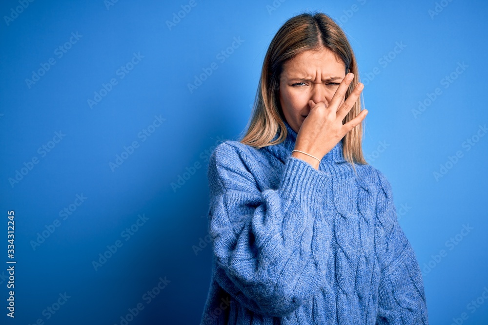 Young beautiful blonde woman wearing winter wool sweater over blue isolated background smelling something stinky and disgusting, intolerable smell, holding breath with fingers on nose. Bad smell Young beautiful blonde woman wearing winter wool sweater over blue isolated background smelling something stinky and disgusting, intolerable smell, holding breath with fingers on nose. Bad smell