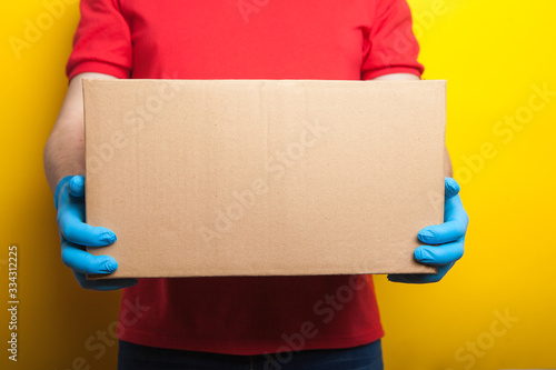 Online ordering and delivery. A man in a red uniform and rubber medical gloves holds a box on a bright yellow background. Food delivery during the coronavirus quarantine period.