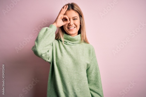 Young beautiful blonde woman wearing winter wool sweater over pink isolated background doing ok gesture with hand smiling, eye looking through fingers with happy face. © Krakenimages.com