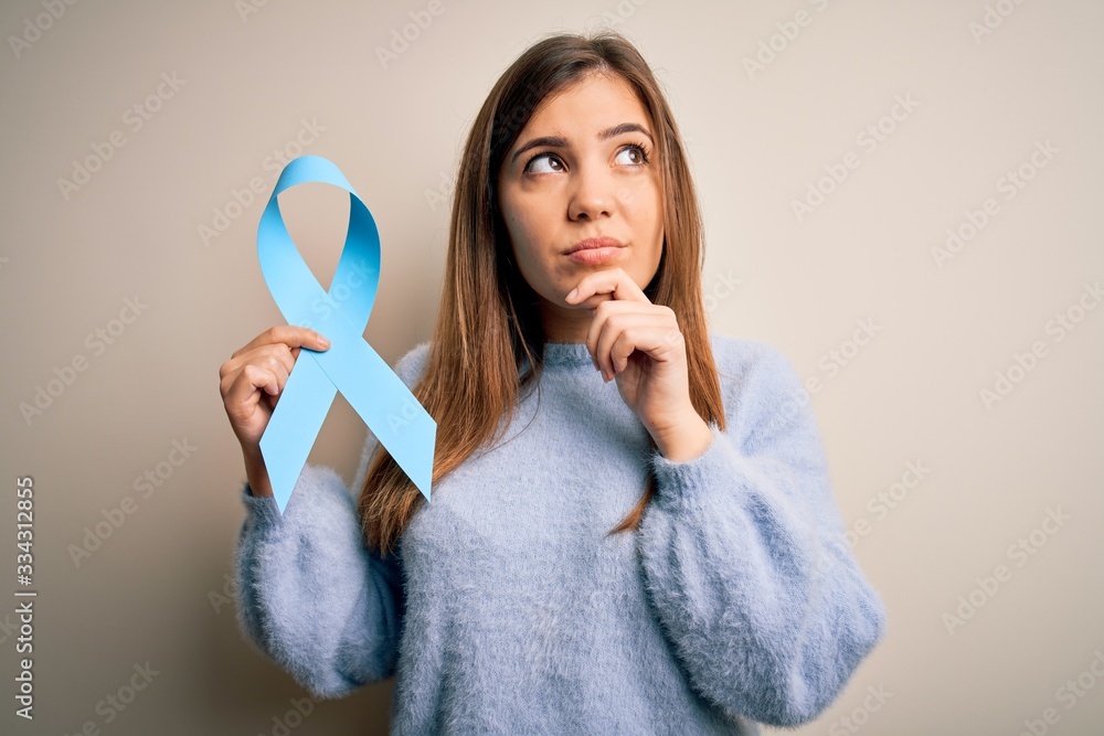 Young blonde woman holding prostate cancer awareness campaing blue ribbon serious face thinking about question, very confused idea Young blonde woman holding prostate cancer awareness campaing blue ribbon serious face thinking about question, very confused idea