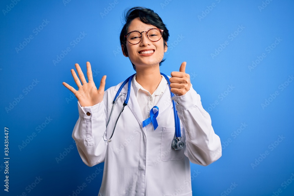 Young beautiful asian doctor girl wearing stethoscope and coat with blue cancer ribbon showing and pointing up with fingers number six while smiling confident and happy. Young beautiful asian doctor girl wearing stethoscope and coat with blue cancer ribbon showing and pointing up with fingers number six while smiling confident and happy.
