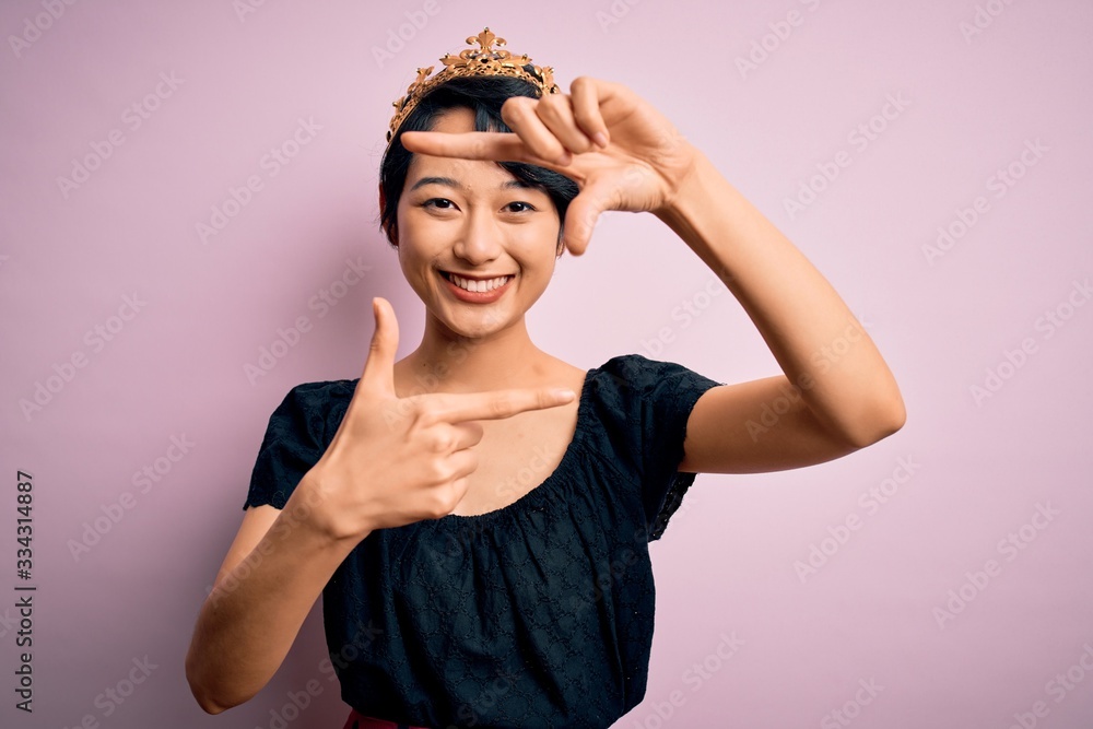 Fototapeta premium Young beautiful chinese woman wearing golden crown of king over isolated pink background smiling making frame with hands and fingers with happy face. Creativity and photography concept.