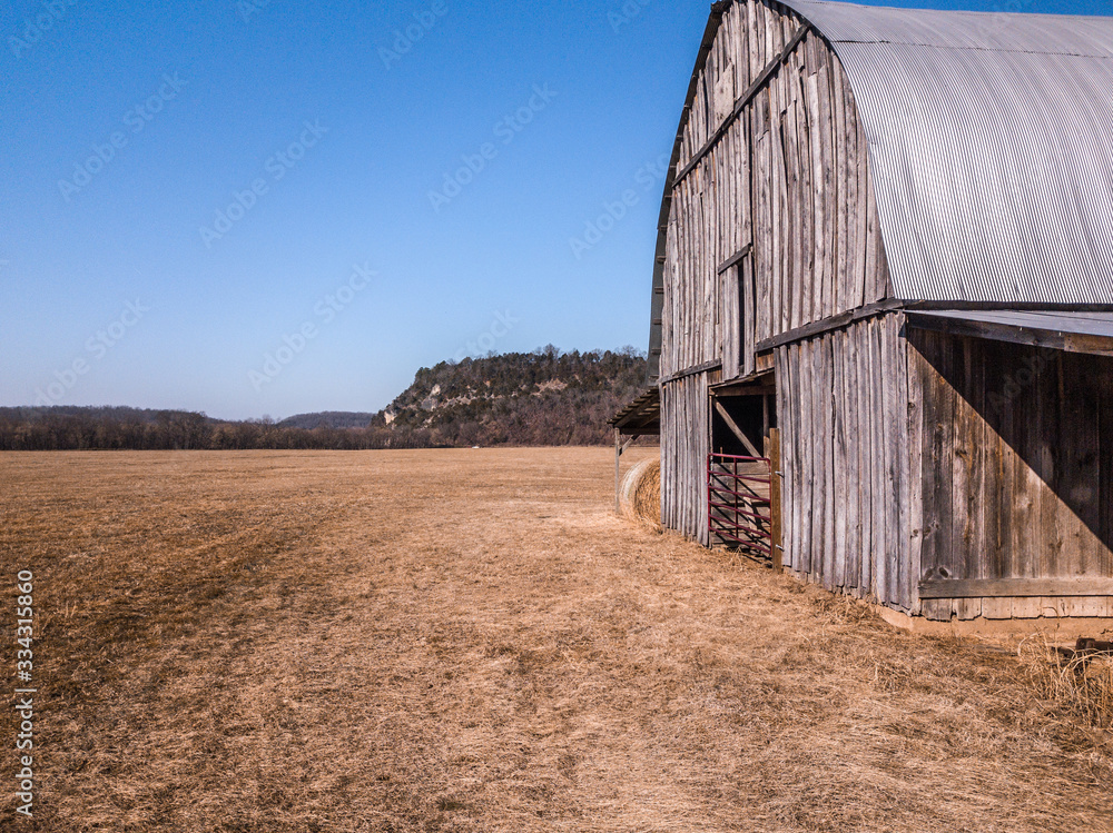 Fototapeta premium Barn in empty valley field by mountain range with hay bales and red baler
