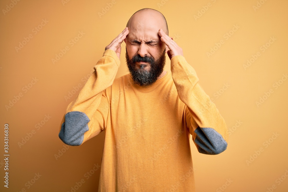 Handsome bald man with beard wearing casual sweater standing over yellow background with hand on head for pain in head because stress. Suffering migraine. Handsome bald man with beard wearing casual sweater standing over yellow background with hand on head for pain in head because stress. Suffering migraine.