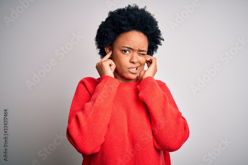 Young beautiful African American afro woman with curly hair wearing red casual sweater covering ears with fingers with annoyed expression for the noise of loud music. Deaf concept.