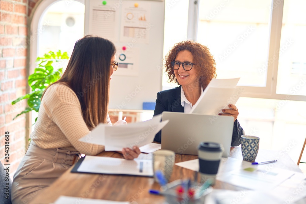 Two beautiful businesswomen smiling happy and confident. Sitting with smile on face working together using laptop at the office Two beautiful businesswomen smiling happy and confident. Sitting with smile on face working together using laptop at the office