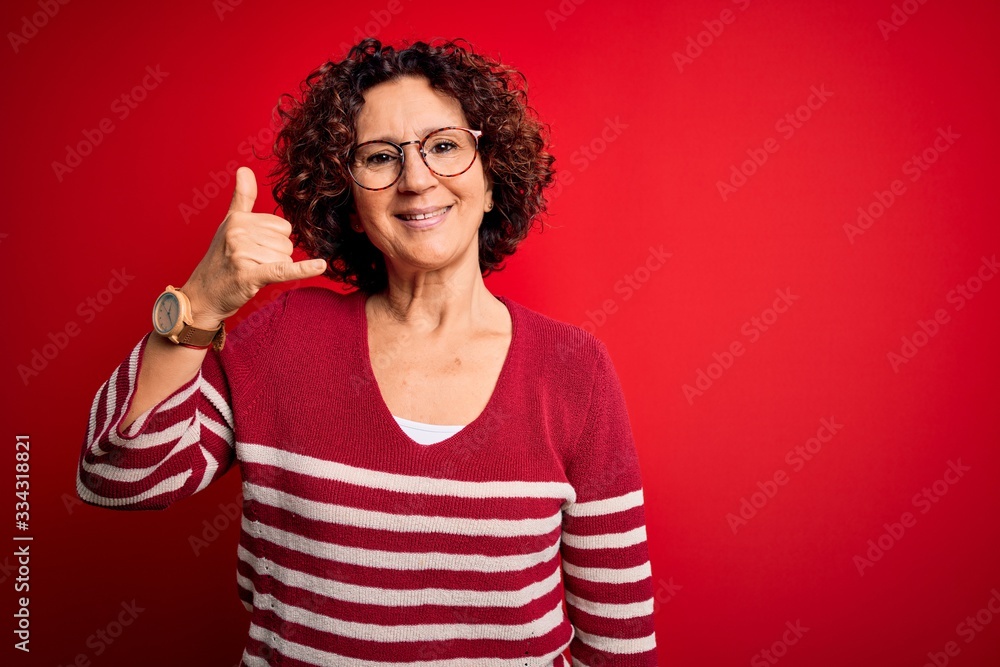 Middle age beautiful curly hair woman wearing casual striped sweater over red background smiling doing phone gesture with hand and fingers like talking on the telephone. Communicating concepts. Middle age beautiful curly hair woman wearing casual striped sweater over red background smiling doing phone gesture with hand and fingers like talking on the telephone. Communicating concepts.