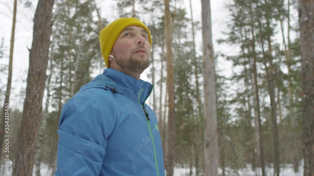 Slowmo tracking with low angle of handsome man with red cheeks standing in forest on cold winter day and looking around while catching his breath after jog Slowmo tracking with low angle of handsome man with red cheeks standing in forest on cold winter day and looking around while catching his breath after jog
