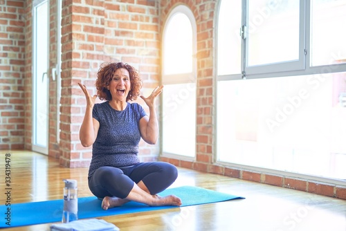 Middle age beautiful sportswoman wearing sportswear sitting on mat practicing yoga at home celebrating crazy and amazed for success with arms raised and open eyes screaming excited. Winner concept