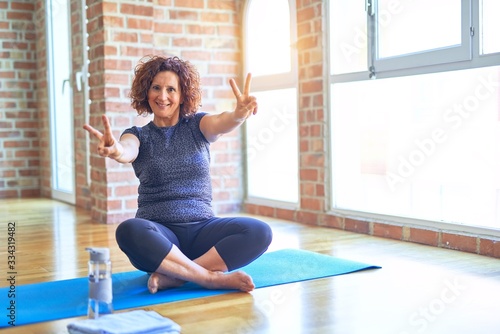Middle age beautiful sportswoman wearing sportswear sitting on mat practicing yoga at home smiling looking to the camera showing fingers doing victory sign. Number two.