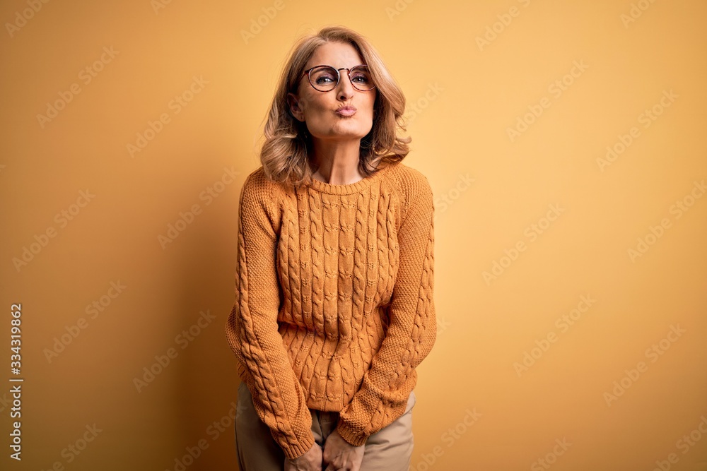 Middle age beautiful blonde woman wearing casual sweater and glasses over yellow background looking at the camera blowing a kiss on air being lovely and sexy. Love expression.