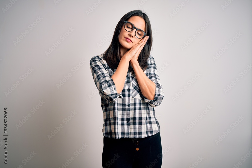 Young brunette woman with blue eyes wearing casual shirt and glasses over white background sleeping tired dreaming and posing with hands together while smiling with closed eyes. Young brunette woman with blue eyes wearing casual shirt and glasses over white background sleeping tired dreaming and posing with hands together while smiling with closed eyes.