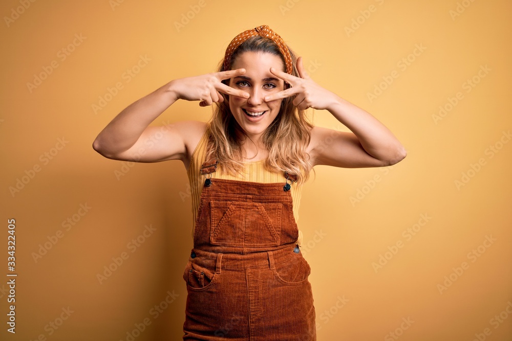 Young beautiful blonde woman wearing overalls and diadem standing over yellow background Doing peace symbol with fingers over face, smiling cheerful showing victory Young beautiful blonde woman wearing overalls and diadem standing over yellow background Doing peace symbol with fingers over face, smiling cheerful showing victory