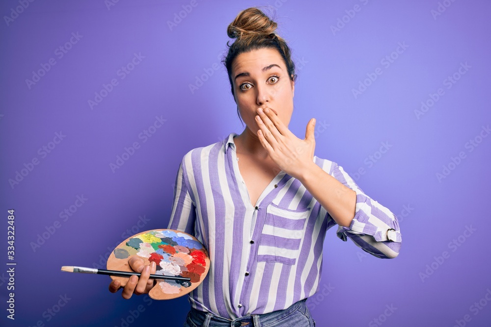 Fototapeta premium Young beautiful blonde artist woman painting using brush and palette over purple background cover mouth with hand shocked with shame for mistake, expression of fear, scared in silence, secret concept
