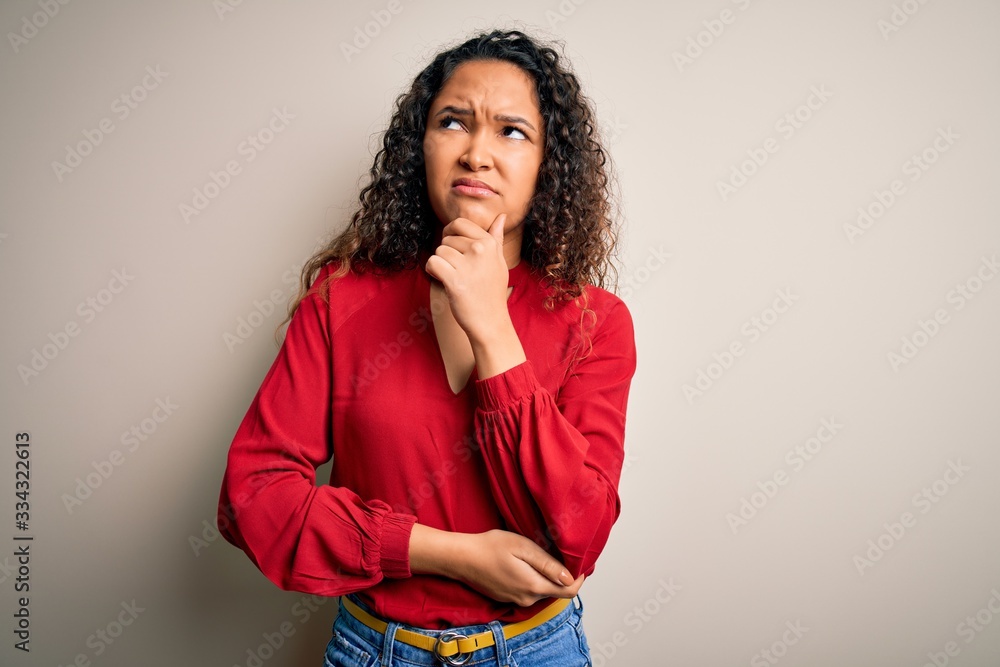 Young beautiful woman with curly hair wearing casual t-shirt standing over white background with hand on chin thinking about question, pensive expression. Smiling with thoughtful face. Doubt concept. Young beautiful woman with curly hair wearing casual t-shirt standing over white background with hand on chin thinking about question, pensive expression. Smiling with thoughtful face. Doubt concept.